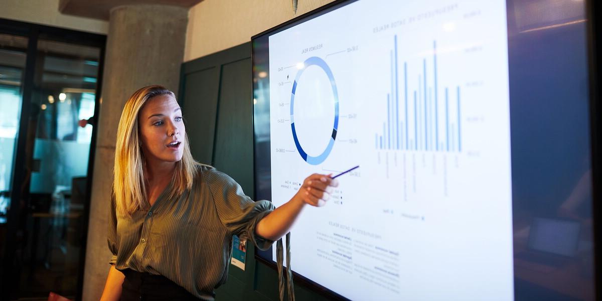 Woman stands and points to a graph on a large monitor while speaking.